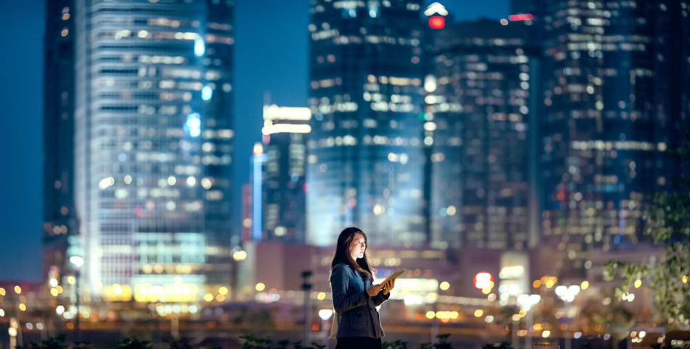 Young businesswoman using digital tablet in financial district, against illuminated corporate skyscrapers at night